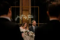 Mariés riants à la table d'honneur, enseigne au néon "just us two" sous une arche dorée au Château Vaudreuil, invités en silhouette au premier plan
