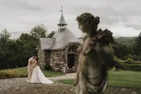 Mariés s'embrassant devant la chapelle de pierre du Château Ste-Agnès, statue en avant-plan flou, ciel nuageux et verdure du domaine viticole