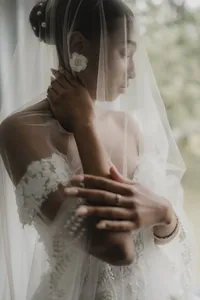 Détail de mariée enveloppée dans son voile, bras levé près du visage, manches de dentelle et bague de fiançailles visibles, lumière douce et tons neutres