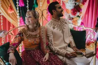 Couple assis côte à côte lors de leur cérémonie du Mehndi, tenues brodées dorées, décor de fleurs colorées et guirlandes en arrière-plan