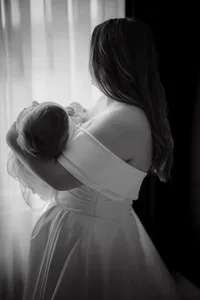 Mariée en robe bustier tenant son bébé blotti contre son épaule, lumière naturelle en contre-jour près d'une fenêtre au Château Vaudreuil, moment de tendresse en noir et blanc