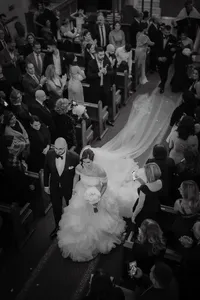 Mariée marchant dans l’allée d’une église aux côtés d’un homme en smoking, entourés de proches émus, sous une lumière douce et poétique, capturant l’intensité et la tendresse du moment en noir et blanc.