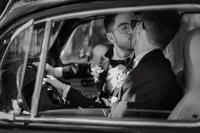 Mariés s'embrassant à bord d'une voiture d'époque, boutonnières florales et nœuds papillon, portrait intime en noir et blanc près du Pont Couvert