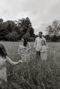A candid black-and-white family photograph of a family of four walking together through tall summer grass in Maine, capturing a natural, timeless moment of connection and togetherness.