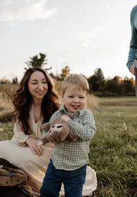 A heartwarming family photo capturing a baby boy running towards the camera with a big smile while mom crouches in the background in a cream dress—joyful, candid lifestyle photography.