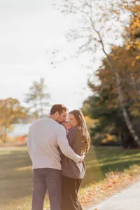 Couple kissing their newborn baby while facing away from the camera, surrounded by warm sunlight with Maine’s beautiful ocean and vibrant fall foliage in the background during a scenic family photography session.