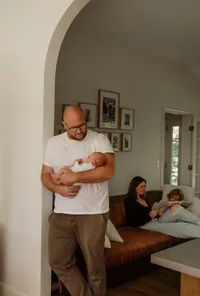 Dad holding their baby in a doorway while mom plays with their older daughter in the background during a Maine family photoshoot — candid New England family photography.