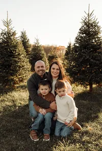 A family kneeling together and smiling during a cozy tree farm photoshoot in Maine, dressed in warm seasonal layers and surrounded by the natural beauty of the evergreens.