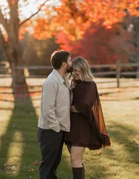 Couple embracing during a peak fall foliage photoshoot in Maine, with the man gently kissing the woman on the forehead as she wears a brown dress and he wears a tan jacket beneath a tree with vibrant autumn leaves.