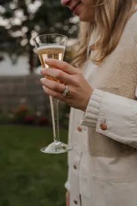 A close-up of an engagement ring on a hand holding a champagne glass, paired with a cream-colored coat for an elegant, timeless detail shot.