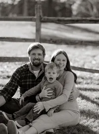 Black and white family photo of a son making a silly face while sitting in the grass with his laughing parents during a fall mini session in Maine, capturing a candid moment of family connection and joy.