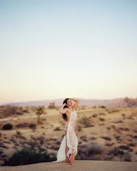 Woman standing barefoot in a desert landscape with sheer white fabric wrapped loosely around her body.