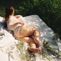 Woman reclining outdoors on layered white blankets in a sheer lace robe, surrounded by grasses and wildflowers.