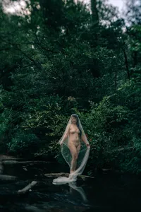 Fine art outdoor boudoir portrait of a woman with sheer fabric surrounded by dense green woods.
