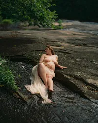 Woman reclining in a shallow stream over smooth rock with pale sheer fabric flowing through the water around her legs.