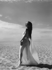 Black-and-white portrait of a woman standing on sand dunes with sheer fabric draped around her body.