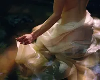 Close-up of a woman seated in shallow water, holding flowing sheer fabric around her body.