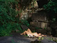 Woman reclining on a stone ledge beside large weathered stone blocks, surrounded by dense green foliage.
