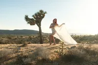 Red-haired woman standing beside a Joshua tree, wrapped in flowing sheer fabric in the sunlit desert.