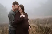 Zwangere vrouw staat met haar rug tegen haar man terwijl ze hun handen vasthouden tijdens een zwangerschapsshoot in de mist in de winter in de omgeving van Apeldoorn