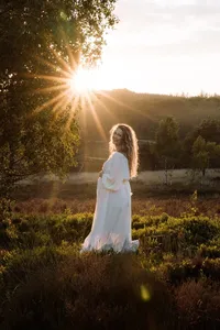 Zwangere vrouw staat met de zon als een ster in haar rug in een witte jurk in de natuur op de veluwe