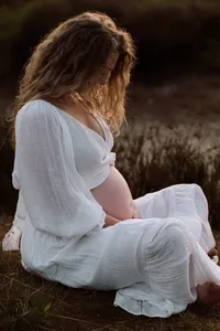 Zwangere vrouw zit in haar witte jurk en haar zwangere buik in de zon in kleermakerszit tijdens een zwangerschapsshoot in de omgeving Apeldoorn
