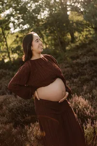 zwanger vrouw kijkt lachend voor zich uit en staat tussen de heide tijdens een zwangerschapsshoot in de omgeving van Apeldoorn