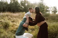 Zwangere moeder geeft haar zoontje een kus terwijl hij op de schouders van zijn vader zit tijdens een fotoshoot op de Veluwe