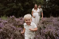 Zoontje rent voor zijn ouders uit tussen de paarse heide en zijn zwangere moeder en vader lopen achter hem aan lachend kijkend naar hun zoon tijdens een zwangerschapsshoot.
