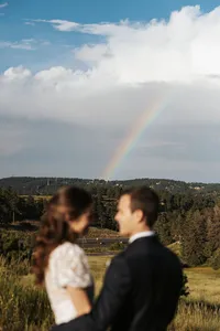A newlywed couple standing close looking at each other with a rainbow in the distance.