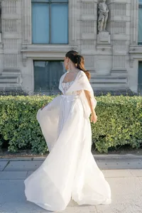 Bride standing alone in a flowing silk gown with lace embroidery captured in motion in a Paris park with historic architecture behind her.