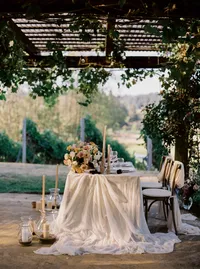 Head table styled with silk linens, garden roses, and ribbon chairs under a vine-covered tent during a Lake Oswego Oregon winery wedding reception.