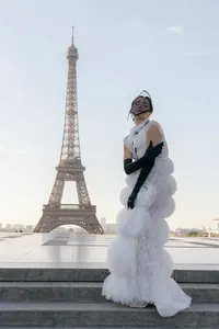 Bride standing in Paris with eyes gently closed, wearing a couture gown with silk and lace details, black gloves, and a structured headpiece near the Eiffel Tower.