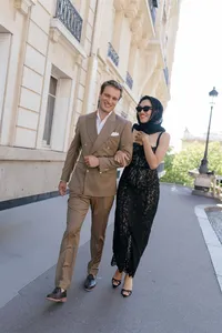 Bride and groom walking through a Paris garden during their engagement session, the bride wearing a black lace dress with gloves and a silk scarf while the groom wears a brown suit.