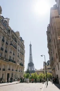 Wide view of the Eiffel Tower rising above Paris gardens and historic buildings with people walking through the park.