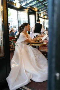 Bride seated at a table at Café de Flore in Paris reading the menu, her voluminous silk gown flowing beside the green café table and burgundy seating in a lively Parisian setting.