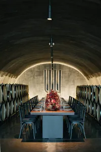 Long rehearsal dinner table styled with deep red florals and elegant glassware inside a winery barrel room.