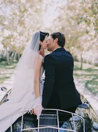 Bride and groom standing at the front of a vintage car, her lace glove resting on the headlight, from a Santa Barbara wedding editorial by Yuliya Supin Design