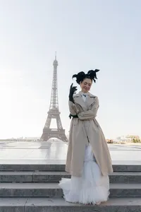 Bride standing in Paris wearing a trench coat and couture gown with the Eiffel Tower softly visible behind her.