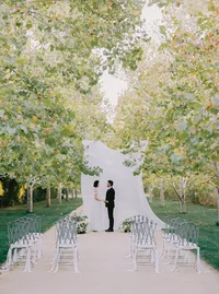 Bride and groom exchanging vows beneath a draped fabric arch along a tree-lined ceremony aisle, from a Santa Barbara wedding editorial by Yuliya Supin Design