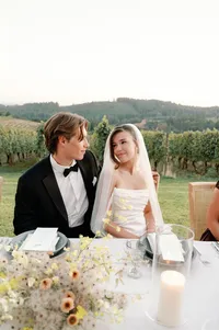 Bride and groom seated at reception table laughing together in warm sunset light.