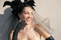 Bride smiling in soft sunlight wearing a couture gown with black gloves and a sheer polka-dot veil headpiece with a statement bow in Paris.