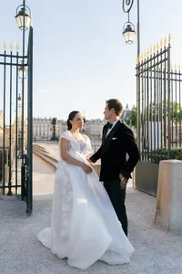 Bride and groom standing together near an iron gate with historic Paris architecture in the background, the bride’s flowing silk and lace gown visible.