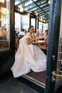 Bride seated at a café table at Café de Flore with her flowing gown extending across the floor, looking toward the camera in the warm café atmosphere.