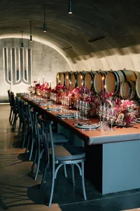 Long table rehearsal dinner design inside a winery cellar surrounded by oak barrels and candlelit floral arrangements.