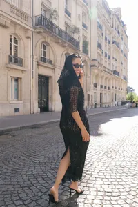 Bride walking along a Paris street during the engagement session wearing a black lace dress, silk scarf, glasses, and red lipstick with historic Paris architecture behind her.