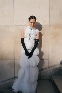 Bride standing against a Paris stone wall wearing a structured couture gown with silk texture, long black gloves, and a statement headpiece.