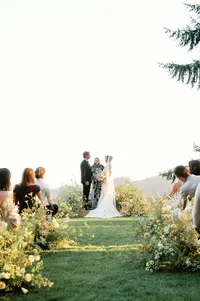 Bride and groom exchanging vows with officiant surrounded by floral-lined aisle and open hills.