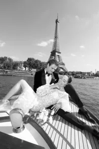Black and white portrait of the bride resting on the groom while holding her bouquet during a boat ride on the Seine River with the Eiffel Tower in the background.