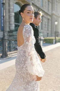 Bride turning toward the camera while walking with the groom through a Paris park with historic buildings in the background.